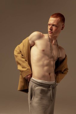 Gri stüdyo arka planında izole edilmiş moda bir kolye takan, düğmesiz sarı gömlekli, şık kızıl saçlı genç adamın portresi. Moda, tarz, beden estetiği, güzellik, erkek sağlığı kavramı