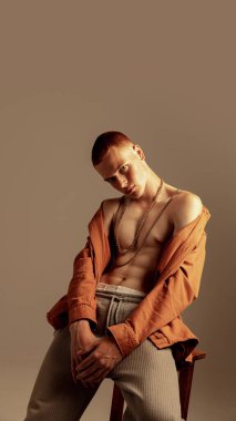 Sandalyeye oturmuş, turuncu gömlekle poz veren, kahverengi arka planda izole edilmiş kameraya bakan genç ve şık kızıl saçlı adamın portresi. Moda, tarz, beden estetiği, güzellik, erkek sağlığı kavramı