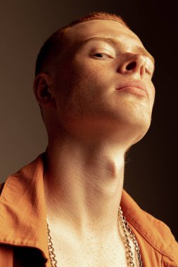 Yüzünde çilleri olan, kahverengi stüdyo arka planında izole edilmiş yakışıklı kızıl saçlı genç adamın yakın plan portresi. Moda, tarz, beden estetiği, güzellik, erkek sağlığı kavramı