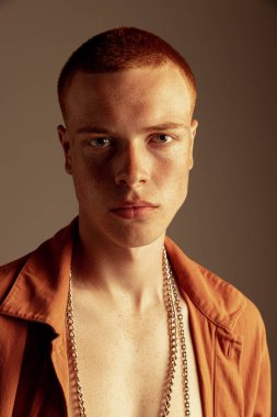 Çilli suratlı yakışıklı kızıl saçlı genç adamın portresi, özenle kahverengi stüdyo arka planında izole edilmiş kameraya bakıyor. Moda, tarz, beden estetiği, güzellik, erkek sağlığı kavramı