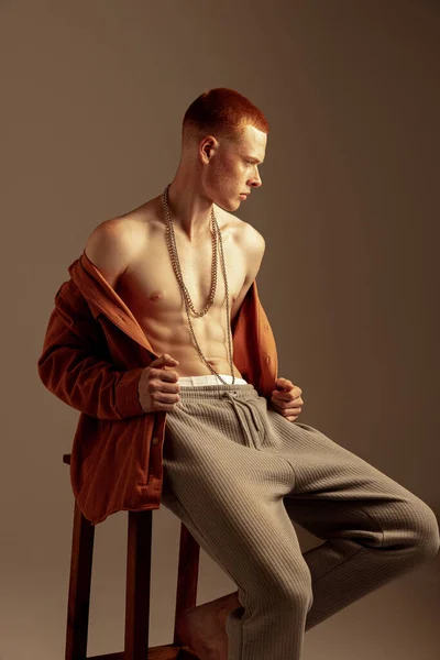 年轻而时尚的红头发男子坐在椅子上 穿着橙色衬衫 与棕色背景隔离 舒缓体形 身体美学 男人健康的概念 — 图库照片