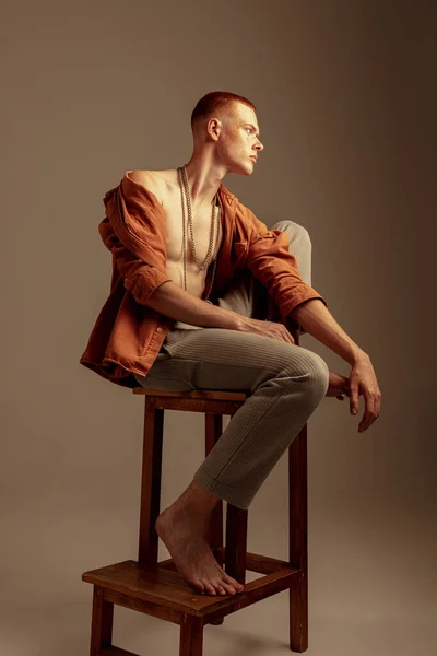 年轻时髦的红头发男孩坐在椅子上的肖像 在棕色的工作室背景上显得孤立无援 身体艺术 身体美学 男性健康 健身的概念 — 图库照片