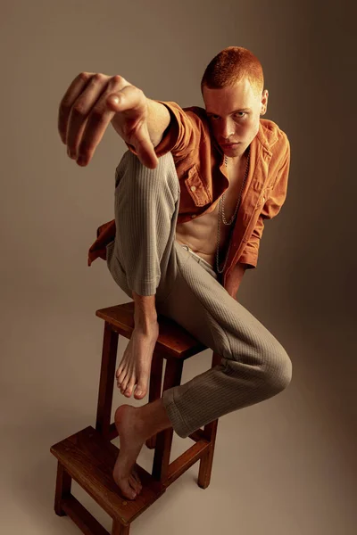 Stylish red-haired young man sitting, posing isolated over brown studio background. Male casual style. Concept of fashion, style, body aesthetics, beauty, mens health. Copy space for ad