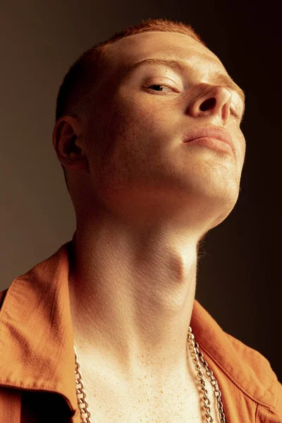 Yüzünde çilleri olan, kahverengi stüdyo arka planında izole edilmiş yakışıklı kızıl saçlı genç adamın yakın plan portresi. Moda, tarz, beden estetiği, güzellik, erkek sağlığı kavramı