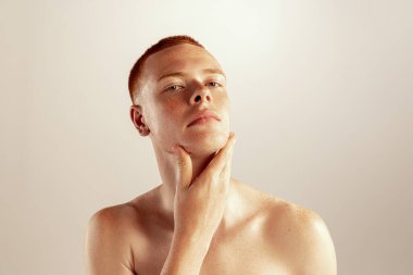 Yanaklarına dokunan çilli kızıl saçlı genç bir adamın portresi. Gri stüdyo arka planında izole bir şekilde poz veriyor. Tıraş makyaj malzemeleri. Erkek sağlığı, yaşam tarzı, güzellik, vücut ve cilt bakımı kavramı