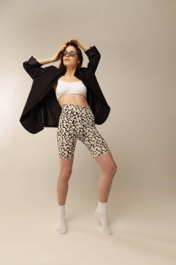 Gri stüdyo arka planında izole edilmiş siyah ceketli ve baskılı taytlı güzel bir kadının tam boy portresi. Tarz kavramı, retro, moda, gençlik, güzellik, duygular