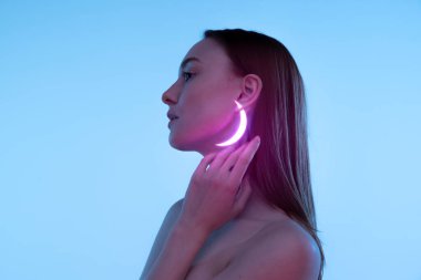 Neon renkli aksesuarları ve mavi arka planda izole edilmiş poz veren küpeleri olan narin genç bir kızın portresi. Mücevher estetiği. Güzellik, yaratıcılık, sanat, sayısallaştırma, moda eğilimleri kavramı