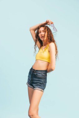 Açık mavi stüdyo arka planında izole edilmiş, günlük kıyafetlerle poz veren kabarık saçlı genç kız portresi. Gençlik, güzellik, yaşam tarzı, moda, eğlence, duygular, tarz kavramı
