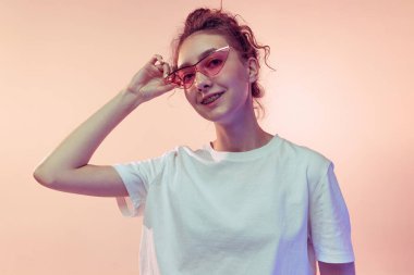 Modaya uygun güneş gözlüğü takan, pembe stüdyo arkaplanından izole edilmiş beyaz tişörtle poz veren şık bir genç kızın portresi. Gençlik kavramı, güzellik, yaşam tarzı, moda, eğlence, duygular