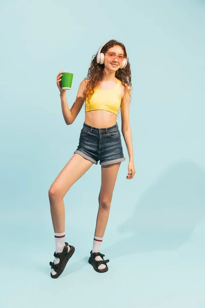 穿着短裤 黄色上衣和耳机的年轻女孩的画像 摆出咖啡杯的姿势 与浅蓝色背景隔离 生活方式 情感的概念 — 图库照片