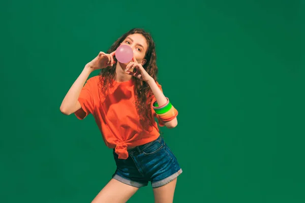 Portrait of cheerful young girl in casual summer outfit, posing with bubble gum isolated on green studio background. Model in orange shirt. Concept of youth, beauty, lifestyle, fashion, fun, emotions