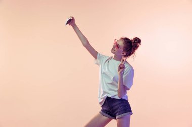 Pembe stüdyo arka planında izole edilmiş cep telefonuyla selfie çeken neşeli genç bir kızın portresi. Modern yaşam tarzı. Gençlik kavramı, güzellik, yaşam tarzı, moda, eğlence, duygular