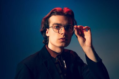 Modaya uygun gözlüklerle poz veren yakışıklı genç adamın portresi, mavi stüdyo arka planında izole edilmiş kameraya bakıyor. Ciddi bir bakış. Gençlik kavramı, moda, yaşam tarzı, duygular. İlan için alanı kopyala