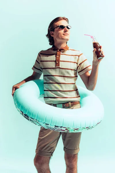 Portrait of young cheerful man in casual outfit and swimming circle, drinking coke, posing isolated over light blue studio background. Concept of youth, fashion, lifestyle, emotions. Copy space for ad