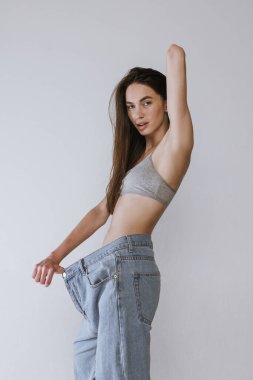 Gri stüdyo arka planında izole edilmiş, büyük boy kot pantolonla poz veren çekici genç kız portresi. Kilo kaybı. Güzellik, vücut ve cilt bakımı, sağlık, kaplıca, kozmetik, reklam