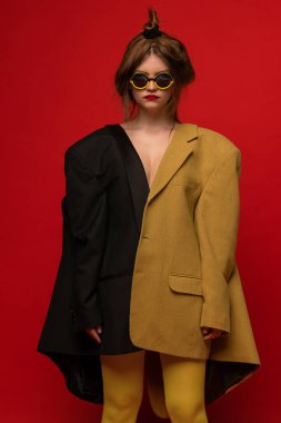Kırmızı arka planda izole edilmiş, iki tane büyük beden ceket, şık, abartılı gözlük takan genç kızın şık portresi. Eski moda kavramı, sanat fotoğrafçılığı, stil, nonoş, güzellik.