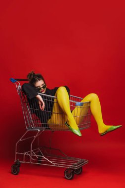 Sarı taytlı genç kızın komik portresi alışveriş arabasında oturmuş, kırmızı arka planda izole bir şekilde poz veriyor. Eski moda kavramı, sanat fotoğrafçılığı, stil, nonoş, güzellik.