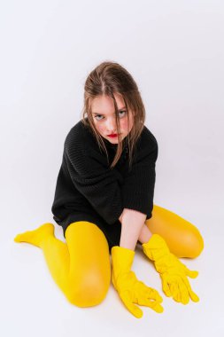 Siyah ceketli, sarı taytlı ve lastik eldivenli genç bir kız, yerde oturuyor, kameraya bakıyor, gri stüdyo arka planında izole bir şekilde poz veriyor. Eski moda kavramı, sanat fotoğrafçılığı, tarz, nonoş.