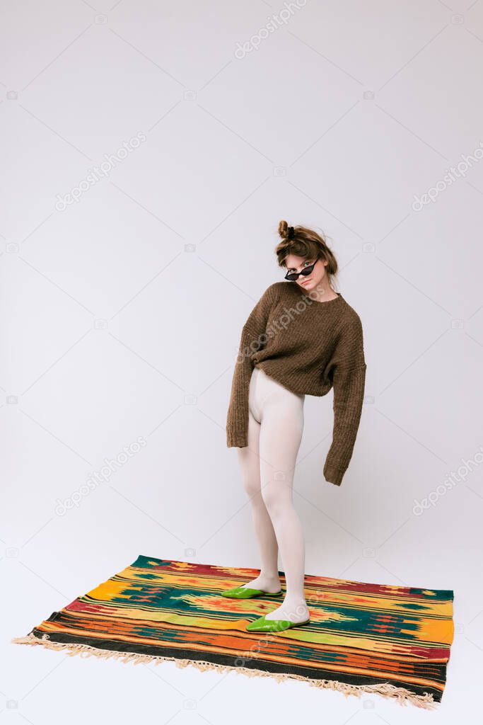 Portrait of stylish young girl in oversized sweater and white tights standing on colorful carpet isolated over grey studio background. Concept of retro fashion, art photography, style, queer, beauty