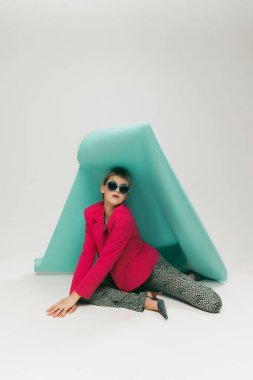 Gri stüdyo arka planında izole edilmiş kağıt parçasıyla poz veren parlak ceketli ve klasik pantolonlu genç kız portresi. Eski moda kavramı, sanat fotoğrafçılığı, stil, nonoş, güzellik.