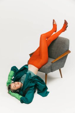 Yeşil ceketli, parlak turuncu dar sandalyeden düşen, gri stüdyo arka planında izole edilmiş şık bir genç kızın portresi. Eski moda kavramı, sanat fotoğrafçılığı, stil, nonoş, güzellik.