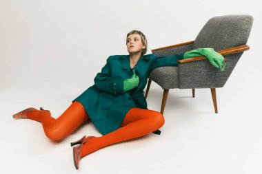 Yeşil ceketli, lastik eldivenli, turuncu taytlı, gri arka planda, sandalyenin yanında poz veren sıra dışı bir genç kız. Eski moda kavramı, sanat fotoğrafçılığı, stil, nonoş, güzellik.