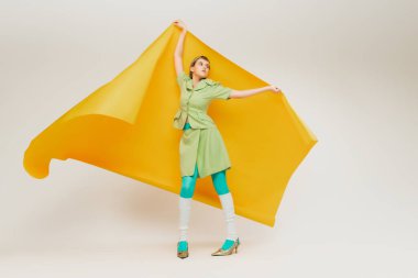 Açık yeşil takım elbiseli şık bir genç kızın portresi, gri stüdyo arka planında izole edilmiş büyük sarı kağıt parçasının altında duruyor. Eski moda kavramı, sanat fotoğrafçılığı, stil, nonoş, güzellik.