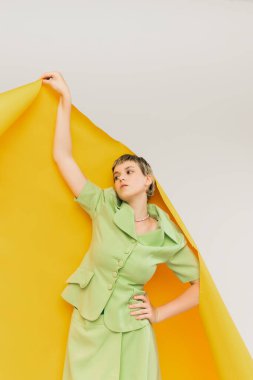 Açık yeşil takım elbiseli şık genç kız, gri stüdyo arka planında izole edilmiş büyük sarı kağıt parçasının altında duruyor. Eski moda kavramı, sanat fotoğrafçılığı, stil, nonoş, güzellik.