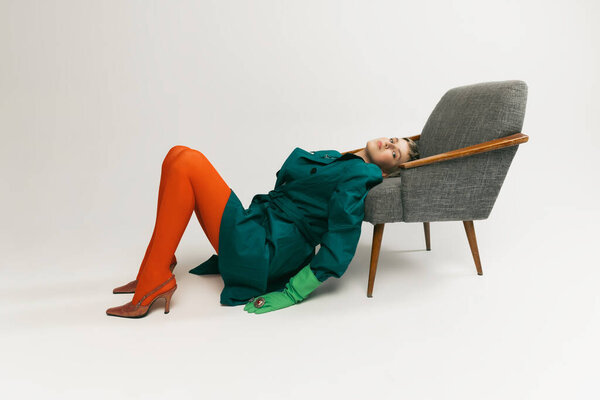 Stylish girl in extraordinary outfit with green coat, rubber gloves and orange tights leaning on chair isolated over grey background. Concept of retro fashion, art photography, style, queer, beauty