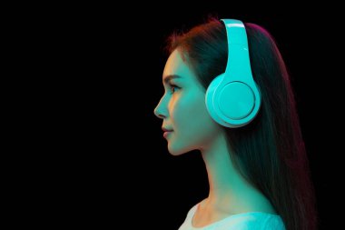 Beyaz kulaklıklarla müzik dinleyen genç bir kızın yan görünüm portresi neon ışıklarıyla izole edilmiş siyah stüdyo arka planında. Güzellik, moda, duygular, yüz ifadesi, gençlik, yaşam tarzı, reklam
