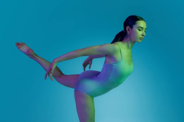 Sıska bir kızın portresi esneme egzersizleri yapıyor mavi stüdyo arka planında neon ışıklarıyla izole edilmiş. Spor bir vücut giysisi giyen bir manken. Spor, sağlık, hobi, gençlik, yaşam tarzı, reklam