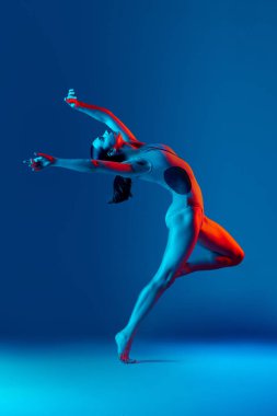 Esneme egzersizleri yapan genç kızın portresi neon ışıklarıyla mavi stüdyo arka planında izole edilmiş. Özgürlüğün atlayışı. Spor, sağlık, hobi, gençlik, yaşam tarzı, reklam
