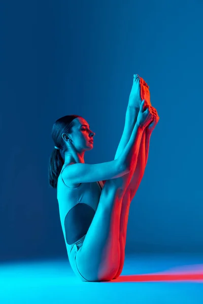 ネオンの光の中で濃い青のスタジオの背景に隔離されたストレッチ練習を行う若いスポーツ少女の肖像画 脚を上げて スポーツ ライフスタイル 広告の概念 — ストック写真