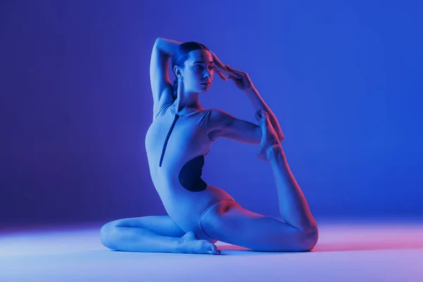 在霓虹灯下 年轻而活泼的女孩在蓝色工作室背景上做伸展腿运动的肖像 业余爱好 生活方式 广告等概念 — 图库照片