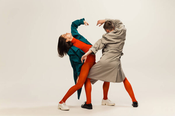 Portrait of stylish man and woman in coats and tights posing isolated over grey studio background. Weirdness, expression. Concept of retro fashion, art photography, style, queer, beauty, party