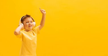 Neşeli küçük bir çocuğun portresi, parlak tişörtlü bir çocuk kulaklıkla müzik dinliyor, sarı arka planda izole edilmiş bir şekilde eğleniyor. Çocukluk kavramı, aile, duygular, yaşam tarzı, moda, reklam