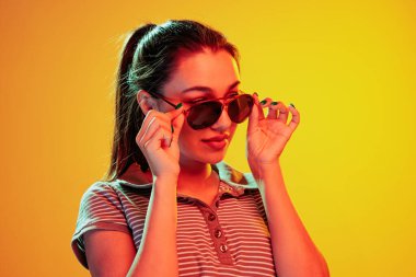 Şık güneş gözlükleriyle poz veren güzel bir genç kızın portresi sarı arka planda, neon ışıkta izole edilmiş. Duyguların kavramı, yüz ifadesi, yaşam tarzı, moda, gençlik kültürü