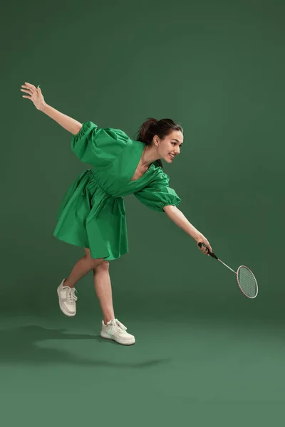 Portrait of cheerful young girl posing in dress, playing badminton isolated over green studio background. Hobby, leisure time. Concept of emotions, facial expression, lifestyle, fashion, youth culture