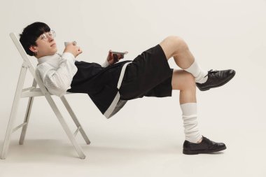 Sandalyeye uzanmış, gri arka planda izole kahve içen şık, genç bir adamın portresi. Siyah beyaz kıyafet. Modern moda kavramı, sanat fotoğrafçılığı, stil, nonoşluk, eşsizlik.