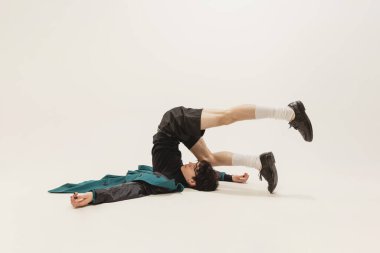 Portrait of stylish young man in black outfit and green coat posing, lying on floor isolated over grey background. Weirdness. Concept of modern fashion, art photography, style, queer, uniqueness, ad