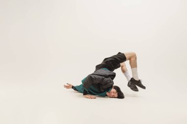 Stylish young man in black outfit and green coat posing, lying on floor isolated over grey studio background. Flexibility. Concept of modern fashion, art photography, style, queer, uniqueness, ad