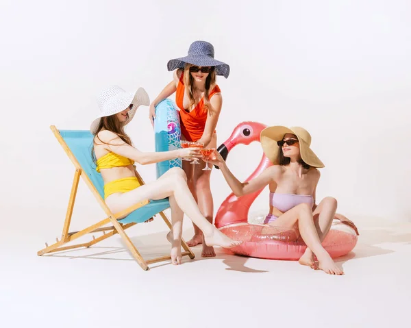 Three cheerful women in swimming suits having summer meeting, swimming, drinking cocktails isolated on grey background. Concept of beauty, fashion, vintage style, summertime, party. Copy space for ad