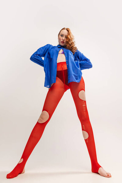 Beautiful young girl in blue shirt and red torn tights posing isolated over grey studio background. Extraordinary, weird look. Concept of retro fashion, art photography, style, queer, beauty, youth