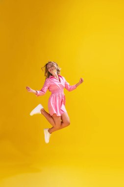 Sarı stüdyo arka planında izole edilmiş şirin pembe elbiseli güzel bir genç kadının portresi. Neşeli atlayışlar. Güzellik kavramı, duygular, yüz ifadesi, yaşam tarzı, moda, gençlik kültürü