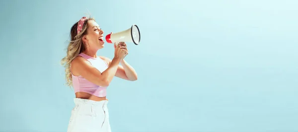 Portrait of young cheerful woman shouting in megaphone isolated over light blue studio background. Summertime sales. Concept of beauty, emotions, facial expression, lifestyle, fashion, youth culture