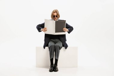 Portrait of stylish young man in total black outfit reading magazine, posing isolated over white studio background. Concept of retro fashion, style, youth culture, emotions, beauty, ad