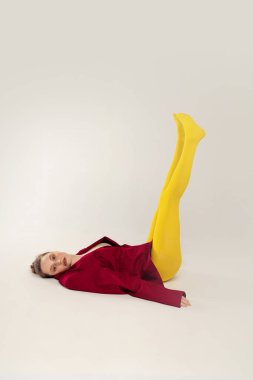 Portrait of young stylish girl in yellow tights and red jacket lying on floor with legs up, posing isolated over grey studio background. Concept of retro fashion, art photography, style, queer, beauty