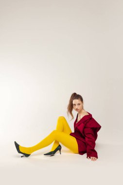 Portrait of stylish young girl sitting, posing in yellow tights, red jacket and heeled shoes isolated over grey studio background. Concept of retro fashion, art photography, style, queer, beauty