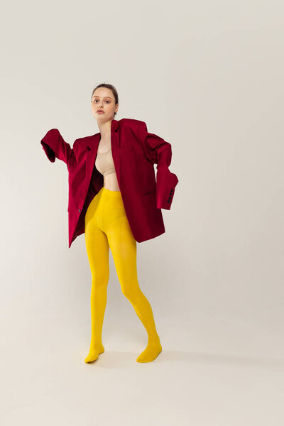 Portrait of young stylish girl in yellow tights and red jacket posing isolated over grey studio background. Extraordinary fashion. Concept of retro fashion, art photography, style, queer, beauty