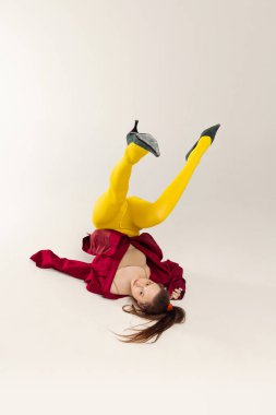 Sarı taytlı, kırmızı ceketli, bacaklarıyla yerde yatan, gri stüdyo arka planında izole edilmiş genç bir kızın portresi. Eski moda kavramı, sanat fotoğrafçılığı, stil, nonoş, güzellik.